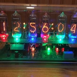 May include: A digital clock with six glowing vacuum tubes displaying the time 2:56:04. The clock has a green circuit board base with colorful LED lights. The tubes are encased in a clear acrylic box.