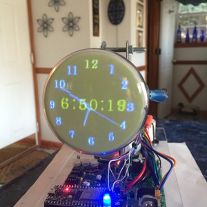 Könnte beinhalten: Eine Digitaluhr mit grünem Display, das die Uhrzeit 6:50:19 anzeigt. Das Zifferblatt ist eine runde Röhre im Vintage-Stil, die auf einer Leiterplatte mit Drähten und Komponenten montiert ist. Die Zahlen sind blau und grün.