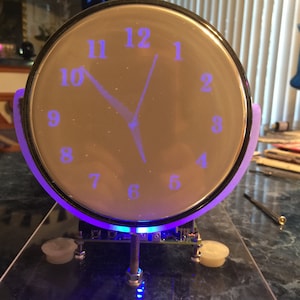 May include: A circular analog clock with a black frame and a translucent face featuring glowing blue numbers from 1 to 12. The clock hands are also illuminated in blue. It is mounted on a clear acrylic base with visible electronic components.