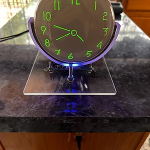 May include: A circular clock with a light gray face and glowing neon green numbers from 1 to 12. The clock features matching green hour and minute hands, mounted on a clear acrylic stand with a blue LED light base and exposed electronic components.