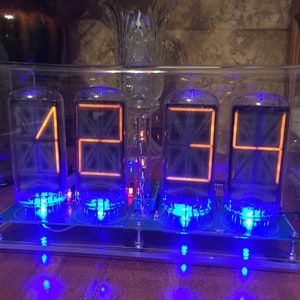 Könnte beinhalten: Ein transparentes Acrylgehäuse beherbergt vier orangefarbene Nixie-Röhren, die die Zeit "12:34" anzeigen. Die Röhren werden mit blauen LEDs beleuchtet.