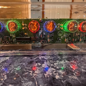 May include: A nixie tube clock featuring six glowing orange numerical digits on a green circuit board. The tubes are illuminated with blue, green, and red LED lights. The device is encased in a clear acrylic housing with visible electronic components.