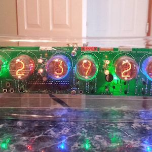 May include: A clear acrylic case encloses a circuit board with six Nixie tubes displaying the time "12:39:22". The tubes glow with a mix of orange, green, and blue light.