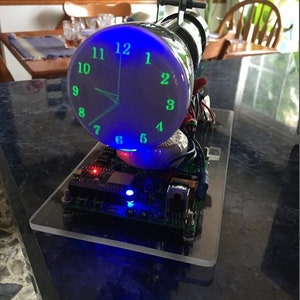 May include: A unique, illuminated clock with a blue display and green numerals. The clock face is a rounded glass tube, mounted on a clear acrylic base with electronic components. The time is displayed with glowing green hands.