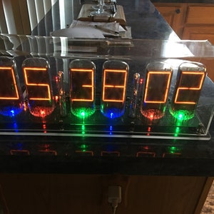 Könnte beinhalten: Eine sechsstellige Nixie-Röhrenuhr, die die Zeit 05:38:02 anzeigt. Die Uhr ist in einem durchsichtigen Acrylgehäuse untergebracht und hat einen schwarzen Sockel. Die Nixie-Röhren leuchten orange, und die Uhr ist mit einem weißen Netzteil angeschlossen.