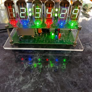 May include: A nixie tube clock featuring six glass vacuum tubes mounted on a green circuit board. The tubes display the numbers 12:34:29 in bright green light, with red and blue LED accents at the base of each tube. The device is supported by metal standoffs.
