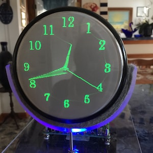 May include: A circular oscilloscope clock featuring a bright green digital display on a dark screen. The clock face shows numbers one through twelve in a stylized font with two glowing green lines indicating the time. It is mounted on a purple frame.