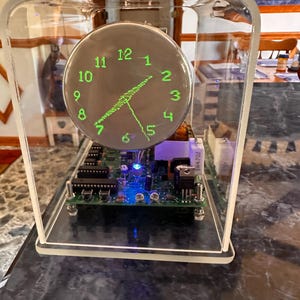 Könnte beinhalten: Eine moderne Uhr in einem transparenten Acrylgehäuse. Das Zifferblatt ist dunkel mit grünen Ziffern und Zeigern. Interne Komponenten, einschließlich einer Leiterplatte, sind sichtbar. Die Uhr steht auf einer dunklen Oberfläche.