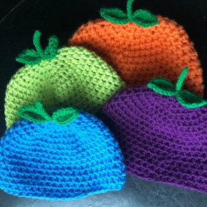 May include: Three crocheted hats in the shape of fruits. The hats are green, orange, and blue. Each hat has a green crocheted leaf on top.