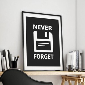 May include: Black and white poster with the text "NEVER FORGET" and a white floppy disk icon on a black background. The poster is framed in black and hanging on a white wall above a wooden desk.