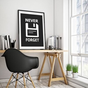 May include: Black and white poster with the text "NEVER FORGET" and a white floppy disk icon on a black background. The poster is framed in black and hanging on a white wall above a wooden desk.