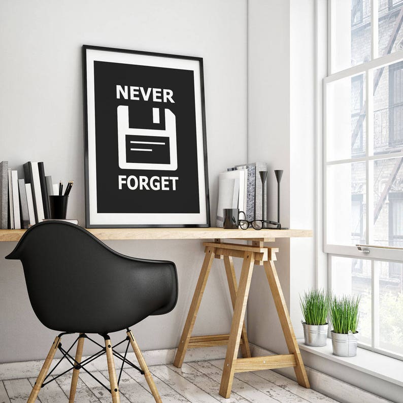 May include: Black and white poster with the text "NEVER FORGET" and a white floppy disk icon on a black background. The poster is framed in black and hanging on a white wall above a wooden desk.