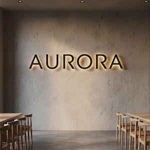 Op de afbeelding: Het woord "AURORA" in zwarte, verlichte letters is bevestigd op een getextureerde, lichtgekleurde muur. De binnenruimte heeft houten tafels en stoelen, wat wijst op een restaurant- of caféomgeving. Het ontwerp is minimalistisch en modern.