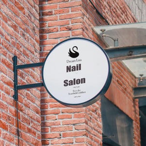 Może przedstawiać: Okrągły, dwustronny szyld salonu paznokci, zamontowany na ceglanej ścianie. Szyld jest biały z czarnym napisem: "Dream Lisa Nail Salon". Czarne logo łabędzia znajduje się na górze. Szyld jest trzymany przez czarny metalowy wspornik.