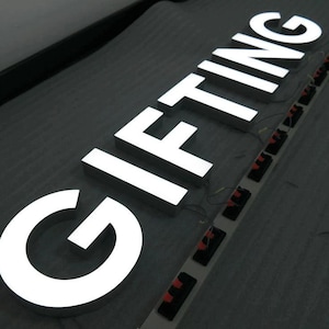 以下が含まれることがあります： 「GIFTING」と綴られた、光る白い金属文字。文字は内側から照らされ、暗い背景に明るい輝きを作り出しています。文字は回路基板に接続されています。