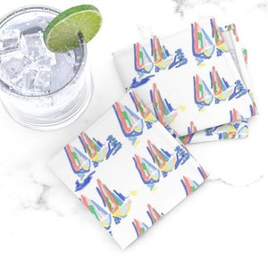 May include: Set of four white cotton napkins with a colorful abstract sailboat design. The napkins are folded and stacked on a white marble surface.
