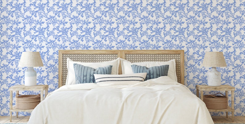 Pode incluir: Um quarto com uma cama branca com almofadas de padr&atilde;o azul e branco e uma cabeceira branca. As paredes s&atilde;o cobertas com um papel de parede floral azul e branco. H&aacute; duas mesas de cabeceira com l&acirc;mpadas e cestos de cada lado da cama.