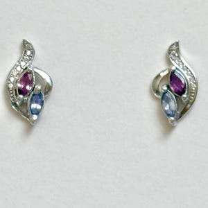 May include: A pair of silver-toned stud earrings featuring a curved, flame-like design. Each earring is set with one marquise-cut purple gemstone and one marquise-cut light blue gemstone, accented by small, clear, sparkling stones along the curve.