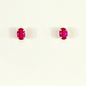 May include: A pair of stud earrings featuring oval-cut, vibrant pinkish-red gemstones set in silver-toned metal prongs. Each gemstone displays a faceted surface that reflects light, creating a clear, polished appearance for these small jewellery pieces.