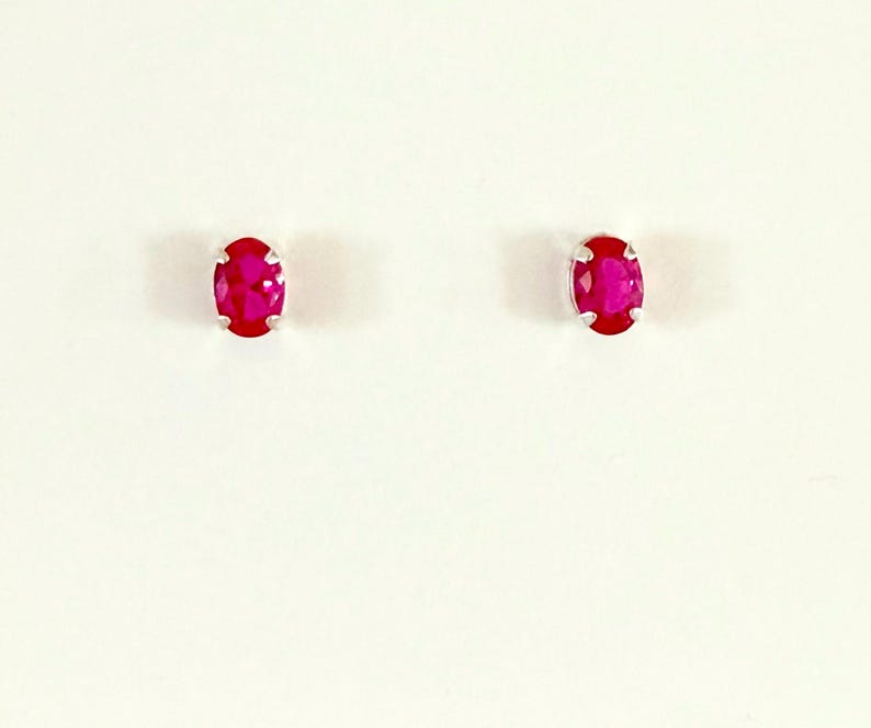May include: A pair of stud earrings featuring oval-cut, vibrant pinkish-red gemstones set in silver-toned metal prongs. Each gemstone displays a faceted surface that reflects light, creating a clear, polished appearance for these small jewellery pieces.