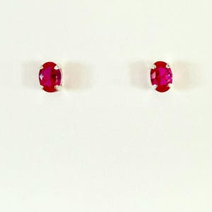 May include: A pair of stud earrings featuring oval-cut, vibrant red gemstones. Each stone is securely held in a four-prong silver-toned metal setting, showcasing a faceted surface that reflects light across the deep crimson coloured faces.