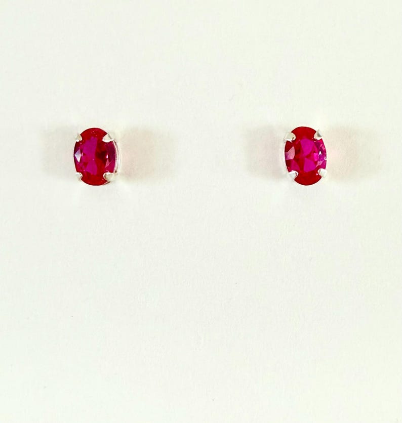 May include: A pair of stud earrings featuring oval-cut, vibrant red gemstones. Each stone is securely held in a four-prong silver-toned metal setting, showcasing a faceted surface that reflects light across the deep crimson coloured faces.