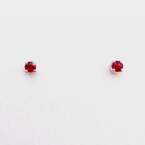 May include: A pair of stud earrings, each featuring a vibrant red gemstone set in a silver-colored metal. The gemstones are round and appear to be securely held in place. The earrings are displayed against a plain white background.