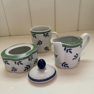 May include: A white ceramic tea set with a green and blue leaf pattern. The set comprises a lidded sugar bowl, a creamer jug, and a mug. The sugar bowl lid has a blue knob. The set is designed for serving tea or coffee.