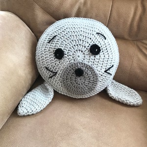 Puede incluir: Un cojín de foca gris tejido a crochet con ojos negros y una nariz negra. El cojín está sentado en un sofá marrón.
