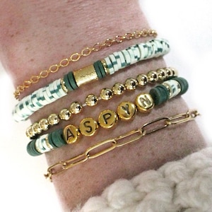 May include: A stack of bracelets with gold chain links, green and white beads, and gold beads with the letters "ASPYN" on them.