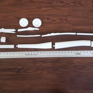 Puede incluir: Un kit de modelo de pl&aacute;stico blanco de un arma futurista, desmontado en varias piezas, dispuesto sobre una superficie de madera. El kit incluye un ca&ntilde;&oacute;n largo y curvo, un mango y varias piezas m&aacute;s peque&ntilde;as.