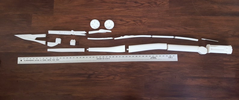 Puede incluir: Un kit de modelo de pl&aacute;stico blanco de un arma futurista, desmontado en varias piezas, dispuesto sobre una superficie de madera. El kit incluye un ca&ntilde;&oacute;n largo y curvo, un mango y varias piezas m&aacute;s peque&ntilde;as.