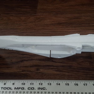 Puede incluir: Un modelo de arma futurista impreso en 3D de color blanco, que se asemeja a una pistola o un rifle futurista. El modelo est&aacute; en dos piezas y est&aacute; sobre una superficie de madera.