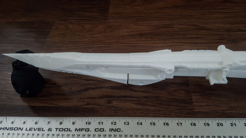 Puede incluir: Un modelo de arma futurista impreso en 3D de color blanco, que se asemeja a una pistola o un rifle futurista. El modelo est&aacute; en dos piezas y est&aacute; sobre una superficie de madera.