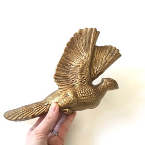 May include: A gold-colored metal wall sculpture of a bird in flight. The bird has detailed feathers and a spread wingspan.