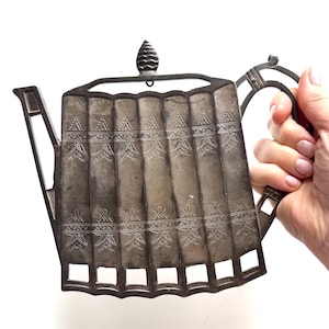 Puede incluir: Una tetera de color plateado con un diseño decorativo grabado. La tetera tiene un cuerpo rectangular con paneles verticales y un asa. La tapa está rematada con un adorno en forma de cono. El pico es angular y el asa es curva.