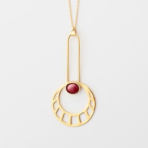 May include: A gold necklace with a geometric pendant featuring a red gemstone. The pendant is a circle with a cut-out design and a vertical bar with a smaller circle at the top.