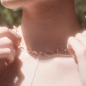 May include: A gold-colored chain necklace with a circular link design. The necklace is being adjusted by a person. The background is blurred, with green foliage and a bouquet of red flowers visible.