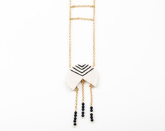 May include: A white ceramic pendant necklace with a black geometric design. The pendant is attached to a gold chain with three gold chains hanging down from the bottom of the pendant. Each of the hanging chains has three black beads.