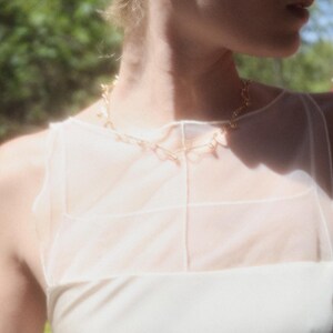 May include: A gold-toned necklace with a delicate design is worn over a white top. The necklace is the focal point, with a simple, elegant style. The top has a sheer overlay, and the background is blurred with greenery and pink flowers.