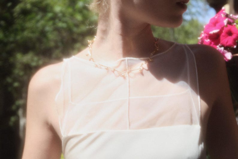 May include: A gold-toned necklace with a delicate design is worn over a white top. The necklace is the focal point, with a simple, elegant style. The top has a sheer overlay, and the background is blurred with greenery and pink flowers.