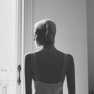 May include: A grayscale image of a person from the back, wearing a light-colored slip dress and a delicate necklace. The person's hair is styled in a classic updo. The scene is illuminated by natural light from a window.