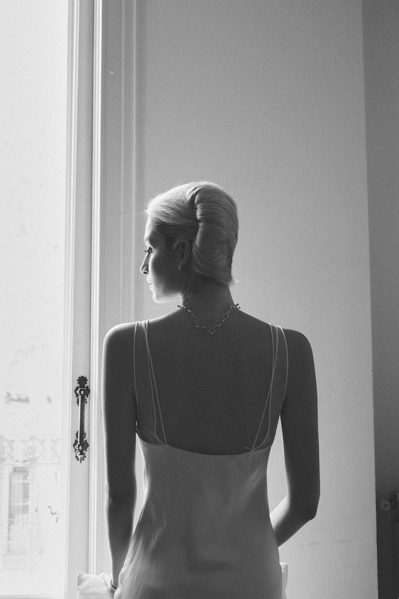May include: A grayscale image of a person from the back, wearing a light-colored slip dress and a delicate necklace. The person's hair is styled in a classic updo. The scene is illuminated by natural light from a window.