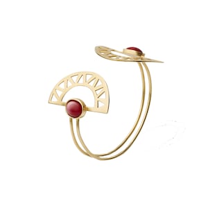 May include: Gold bangle bracelet with two red gemstone accents. The bracelet features a geometric design with a sunburst pattern.
