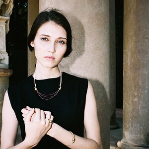 May include: A woman wearing a black sleeveless dress with a gold necklace and bracelet. The necklace has small purple beads and the bracelet has two gold spheres.