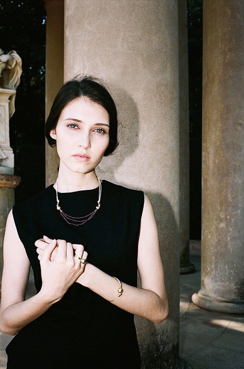 May include: A woman wearing a black sleeveless dress with a gold necklace and bracelet. The necklace has small purple beads and the bracelet has two gold spheres.