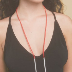 May include: A silver rectangle pendant necklace with a red cord. The necklace is worn by a person with long, dark hair.