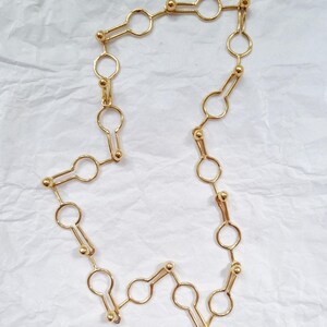 May include: A gold-colored necklace with a unique keyhole-shaped link design. The necklace is made of small, circular links connected by elongated keyhole shapes and small gold beads. The jewelry is displayed on a white, textured surface.