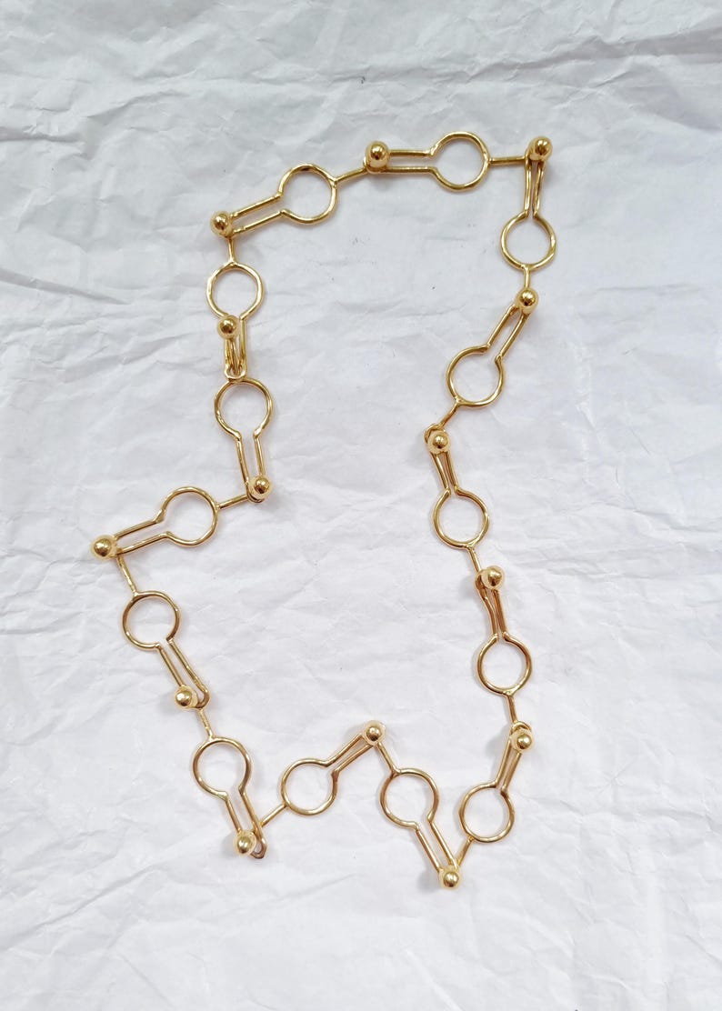 May include: A gold-colored necklace with a unique keyhole-shaped link design. The necklace is made of small, circular links connected by elongated keyhole shapes and small gold beads. The jewelry is displayed on a white, textured surface.