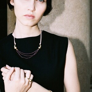 May include: A woman wearing a black sleeveless dress with a gold necklace and bracelet. The necklace has a pink cord and small gold beads. The bracelet is a simple gold band with two small gold balls.